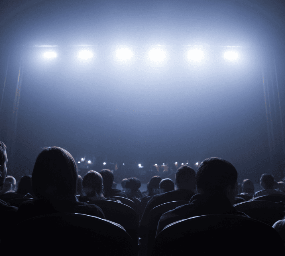 Audience seated in a dimly lit theater, facing a bright stage with overhead lights shining down. - Home Instead