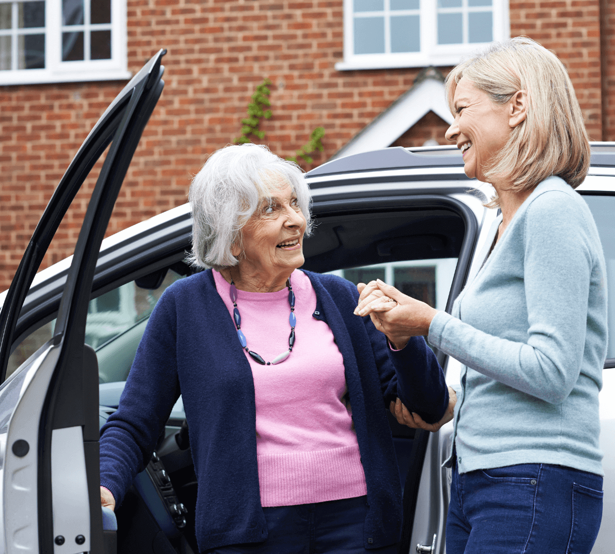 An elderly woman, assisted by a younger woman, steps out of a car in front of a brick house. They are smiling and holding hands. - Home Instead