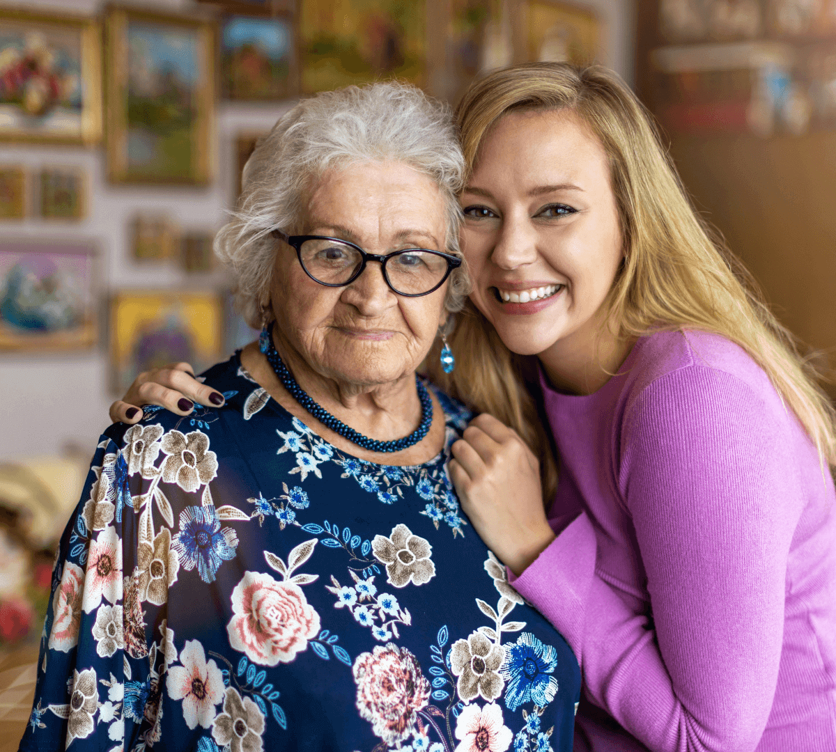 An elderly woman with white hair and glasses poses with a smiling young woman in a purple sweater. - Home Instead