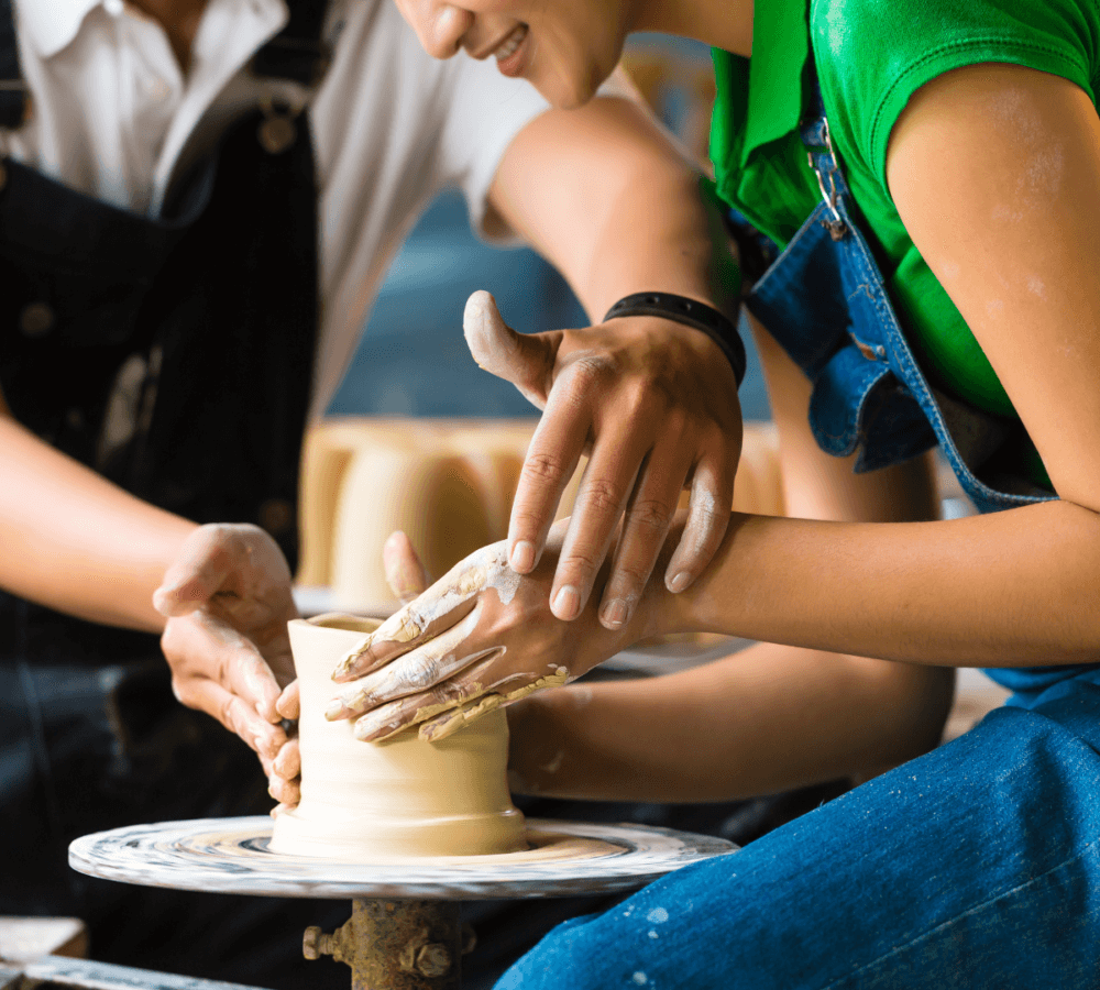 Two people shaping clay on a pottery wheel, hands covered in clay, one guiding the other in a close, collaborative effort. - Home Instead