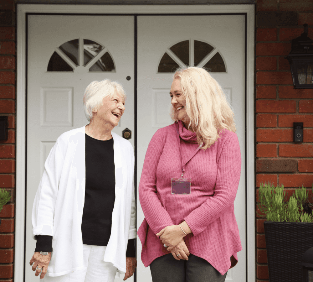 Two women, one elderly and one middle-aged, smiling and talking while standing by a door with potted plants on either side. - Home Instead