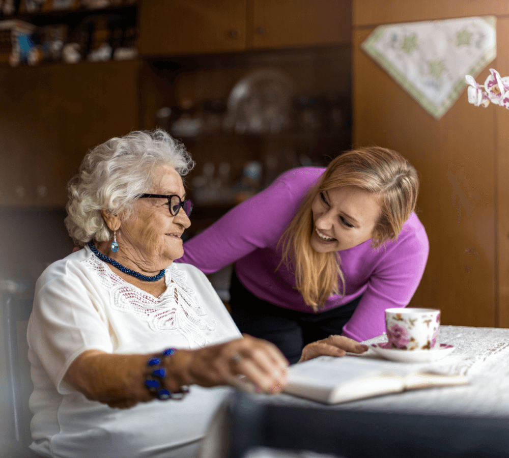 A woman smiles while standing next to an elderly woman who is seated and reading a book at a table with a teacup. - Home Instead