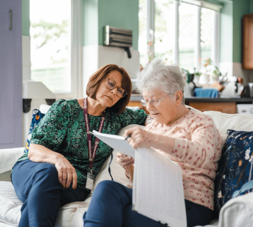 A caregiver and an elderly woman sit on a couch, reviewing a document together in a bright, cozy room. - Home Instead