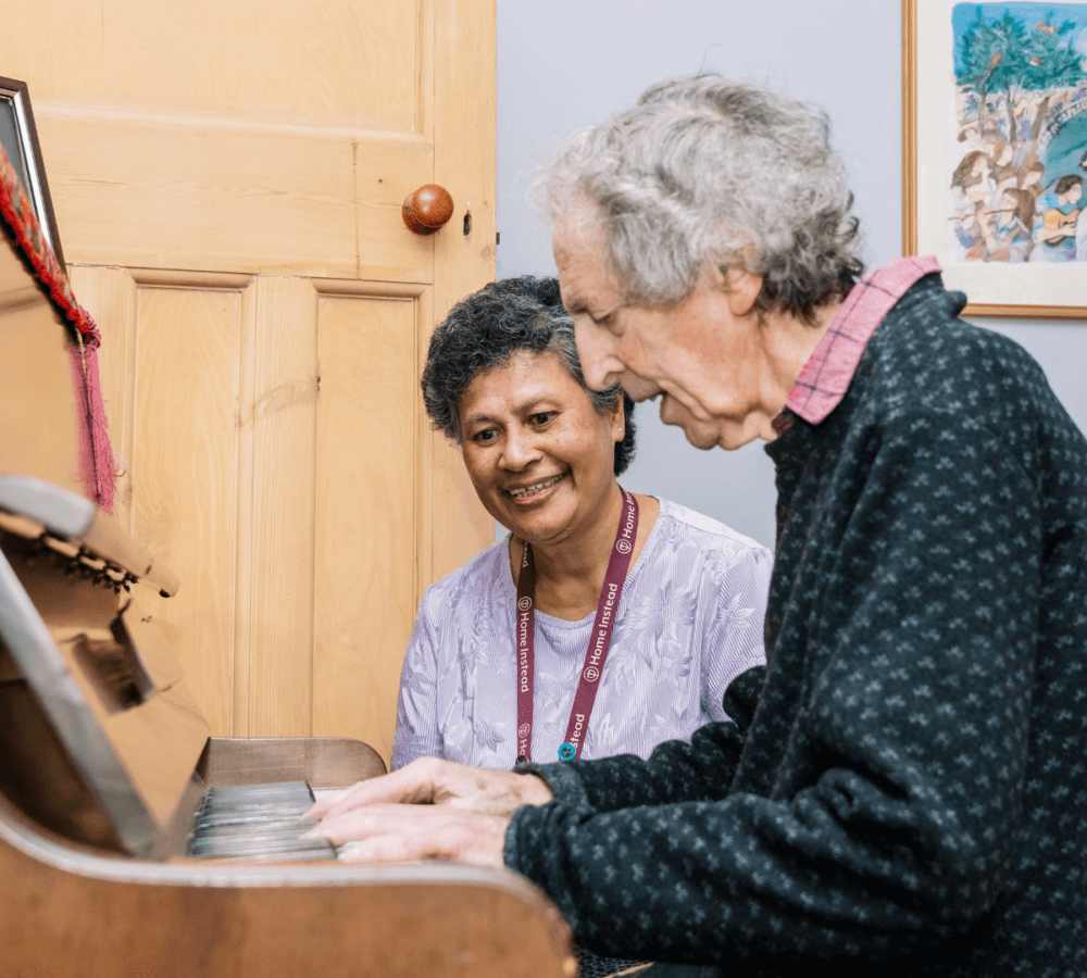 A man plays the piano while a woman watches and smiles in a cozy room. - Home Instead