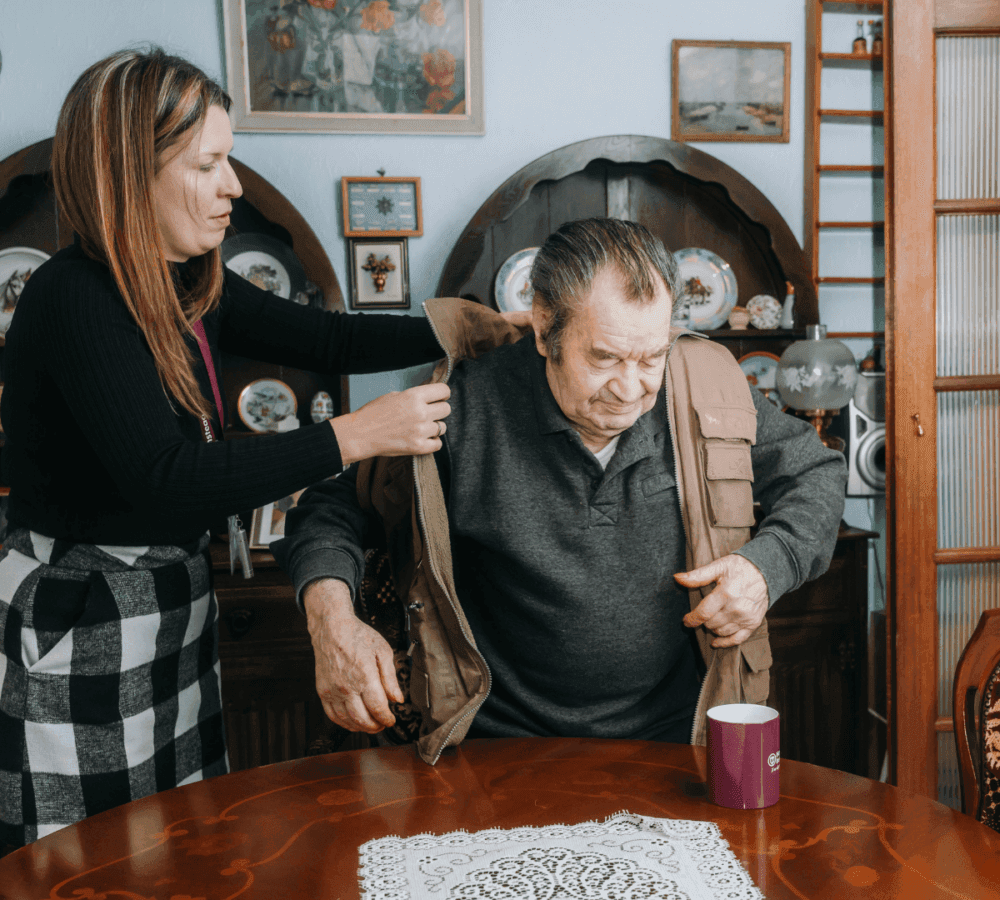 A woman helps an older man put on a jacket at a wooden table with a lace cloth and purple mug in a cozy room. - Home Instead