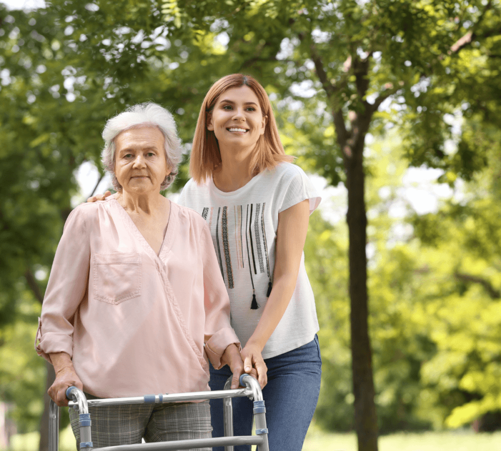 An elderly woman with a walker and a young woman stand together outside, smiling on a sunny day. - Home Instead