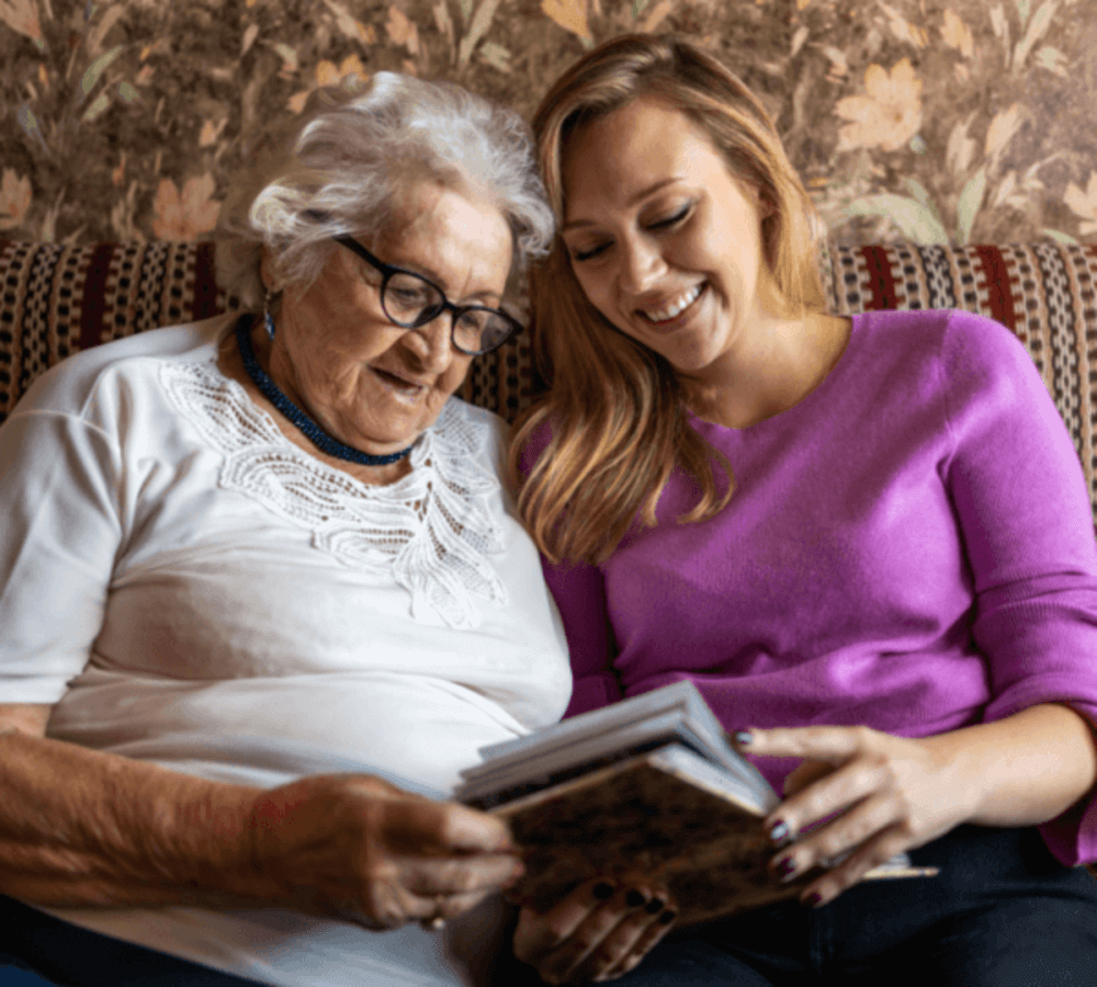 An elderly woman and a younger woman sitting on a couch, smiling while looking at a photo album together. - Home Instead
