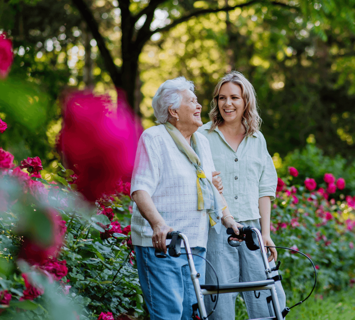 An elderly woman with a walker is smiling while standing next to a younger woman in a garden filled with blooming roses. - Home Instead