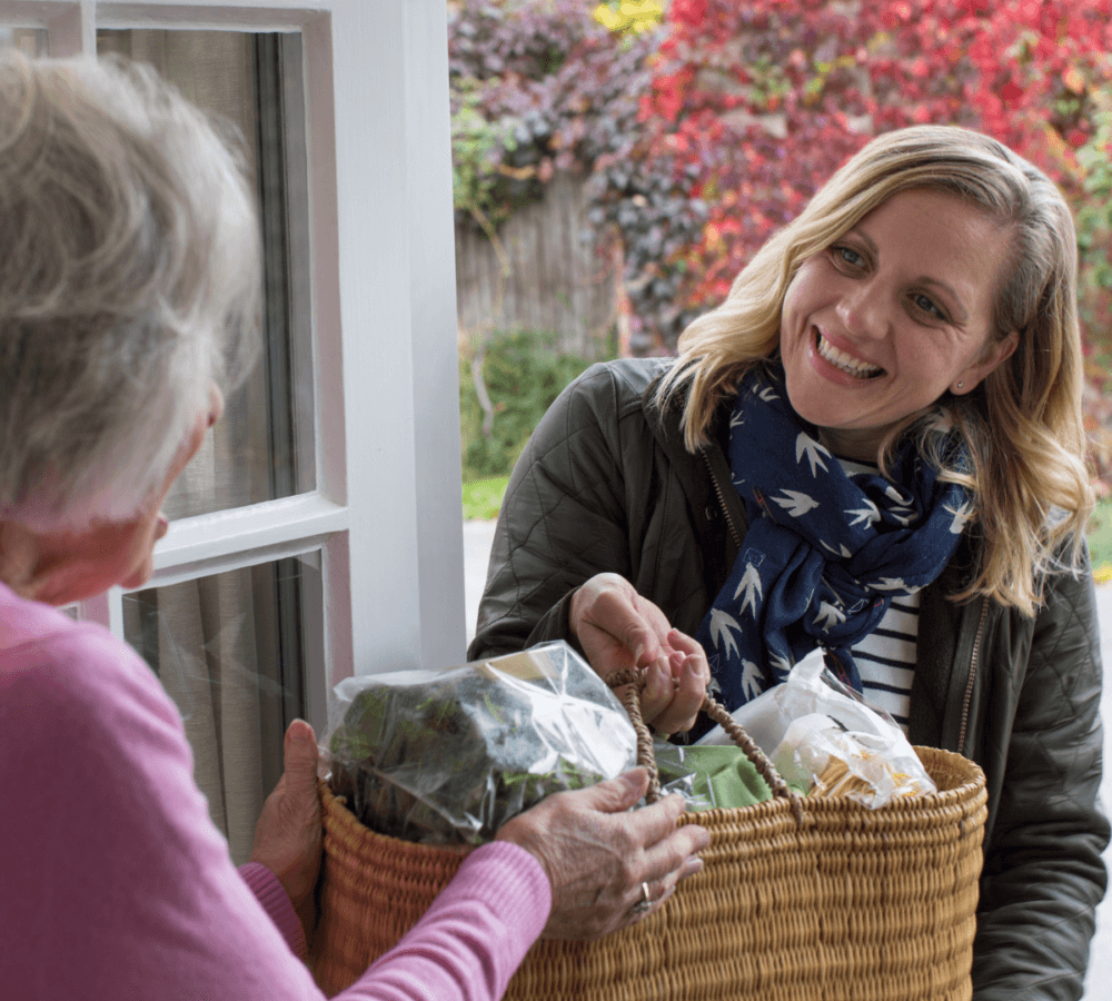 A smiling woman hands a basket of groceries to an elderly woman at the door, with autumn foliage in the background. - Home Instead