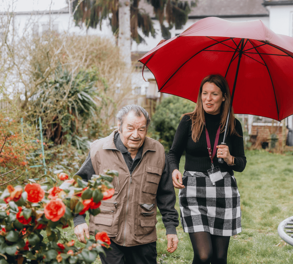 Woman holding a red umbrella walks with an elderly man through a garden with blooming flowers. - Home Instead