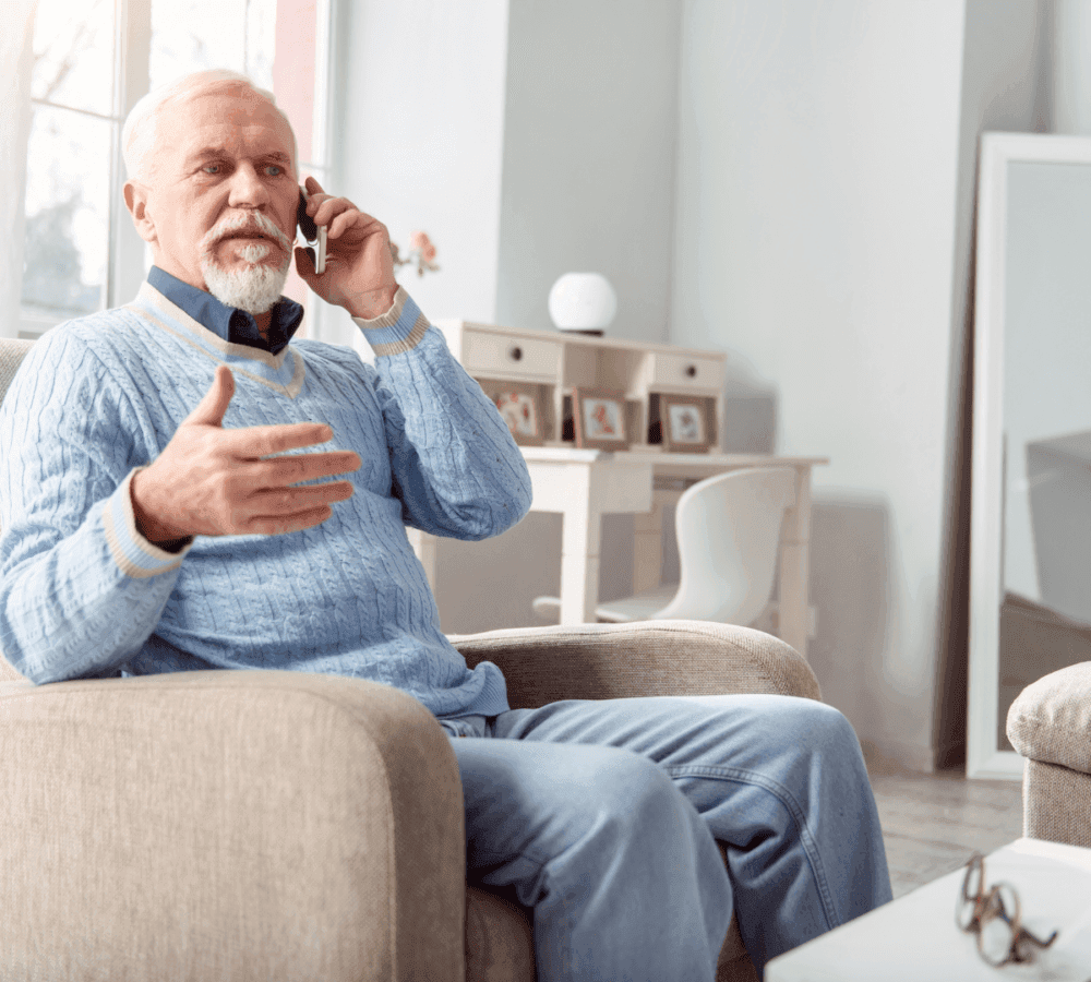 Elderly man with white beard talking on a phone while sitting in a cozy, modern living room. - Home Instead