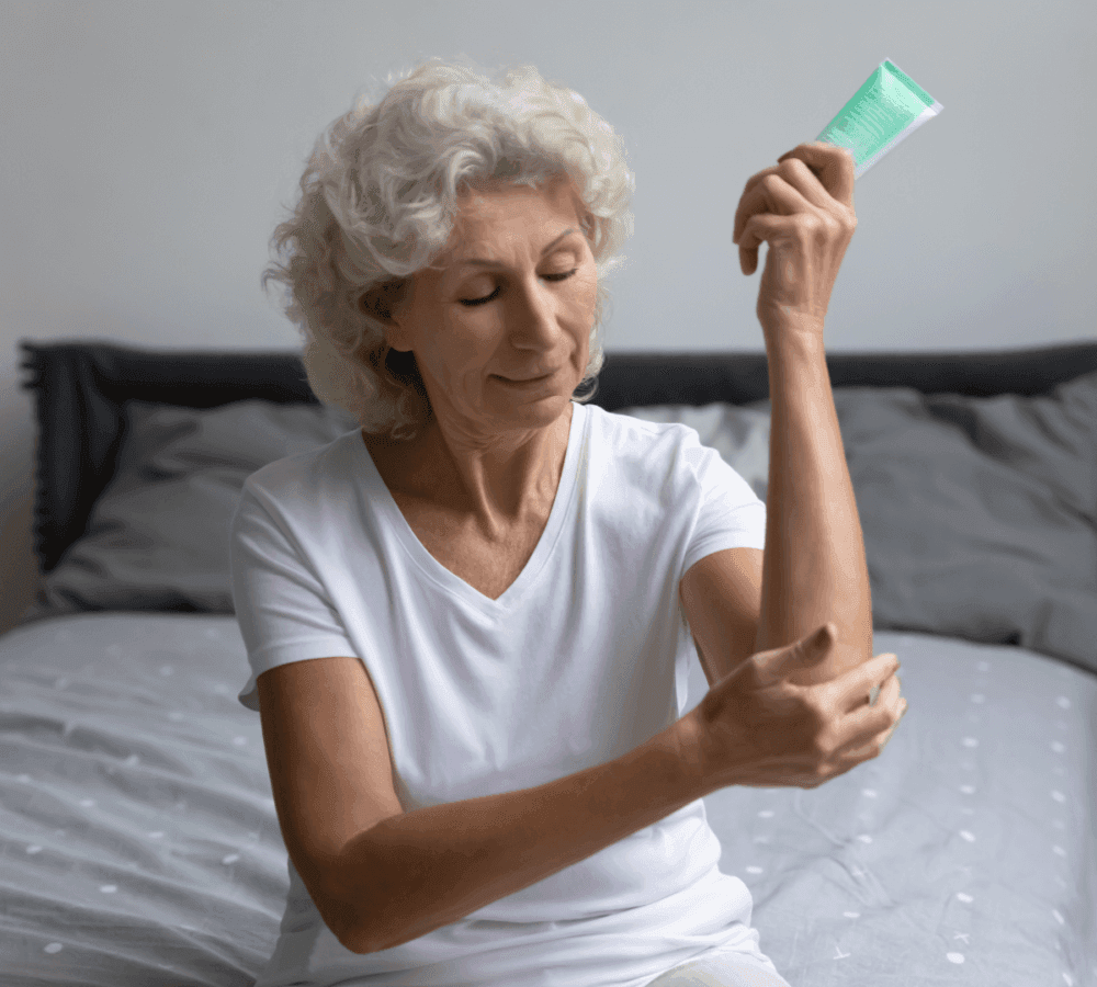 Elderly woman with short curly hair applying lotion to her arm while sitting on a bed, wearing a white shirt. - Home Instead