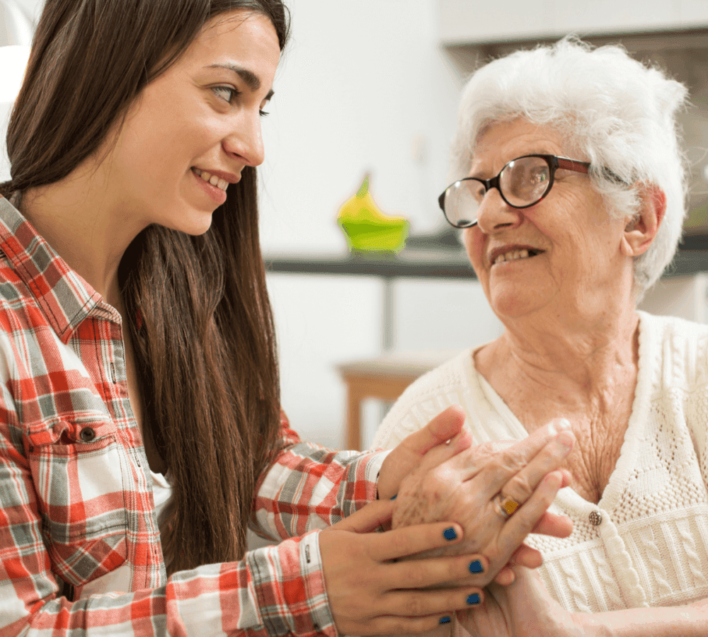 A young woman with long hair holds hands and smiles at an elderly woman wearing glasses and a white sweater. - Home Instead