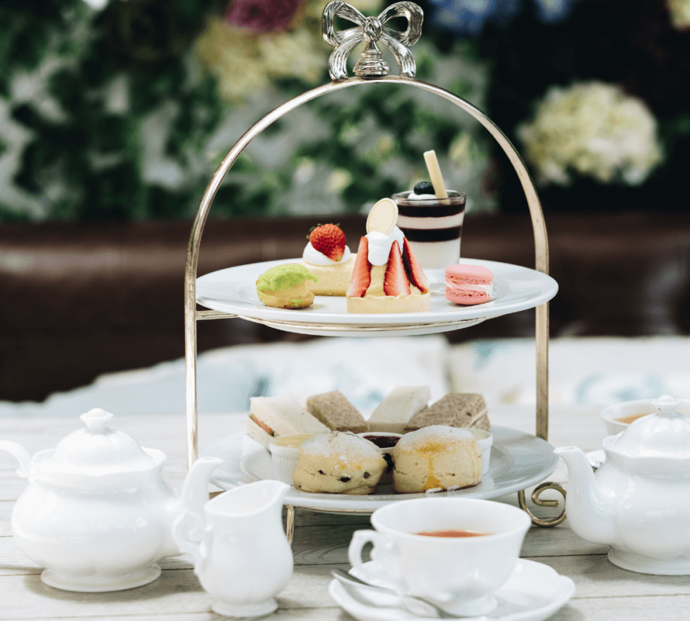 A set of white ceramic tea pots and cups alongside a two-tiered tray with assorted pastries and desserts. - Home Instead