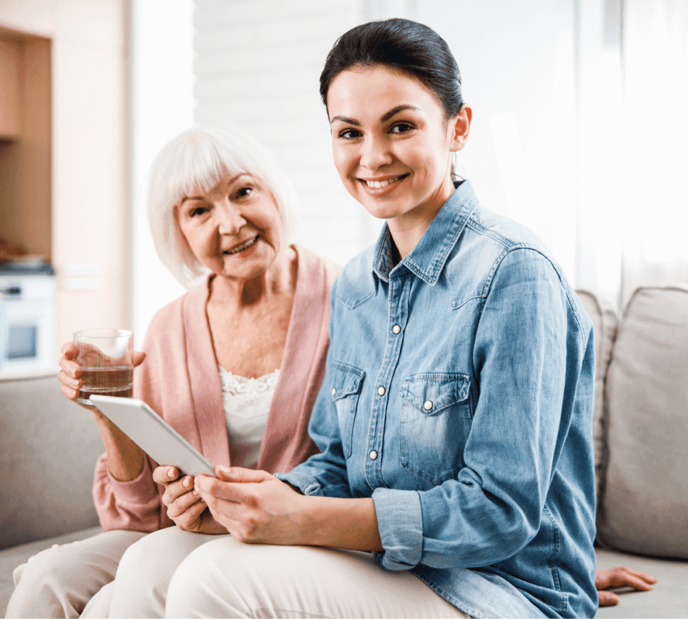 A young woman with a tablet sits next to an elderly woman holding a glass of water, both smiling in a cozy room. - Home Instead