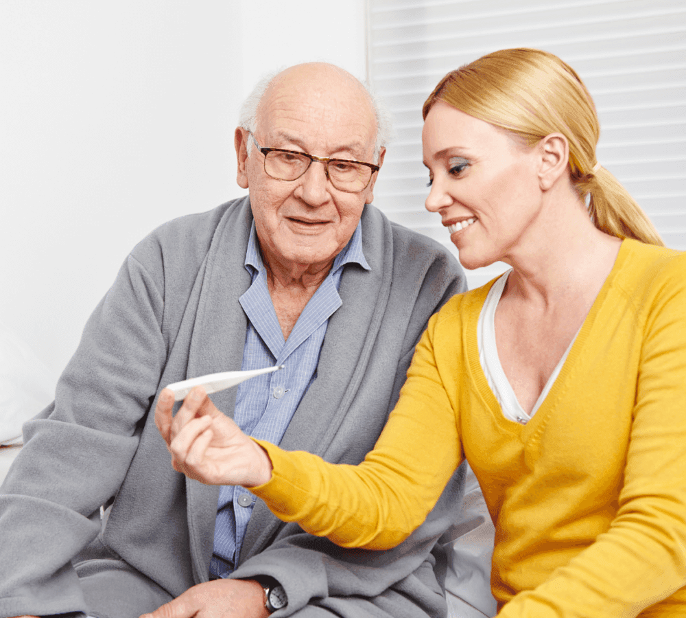 An elderly man and a smiling woman look at a thermometer while sitting together in a bright room. - Home Instead