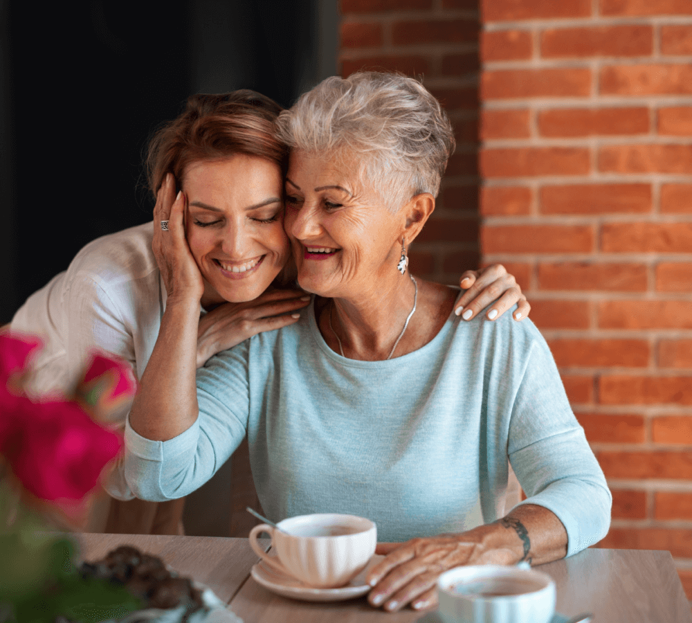 Two women share a loving hug while sitting at a table with coffee cups, with a brick wall in the background. - Home Instead