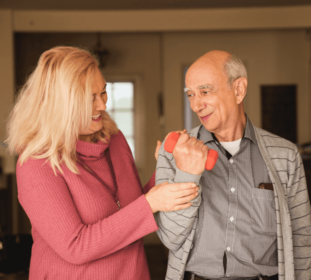 A woman helps an elderly man exercise with a dumbbell in a cozy indoor setting. - Home Instead