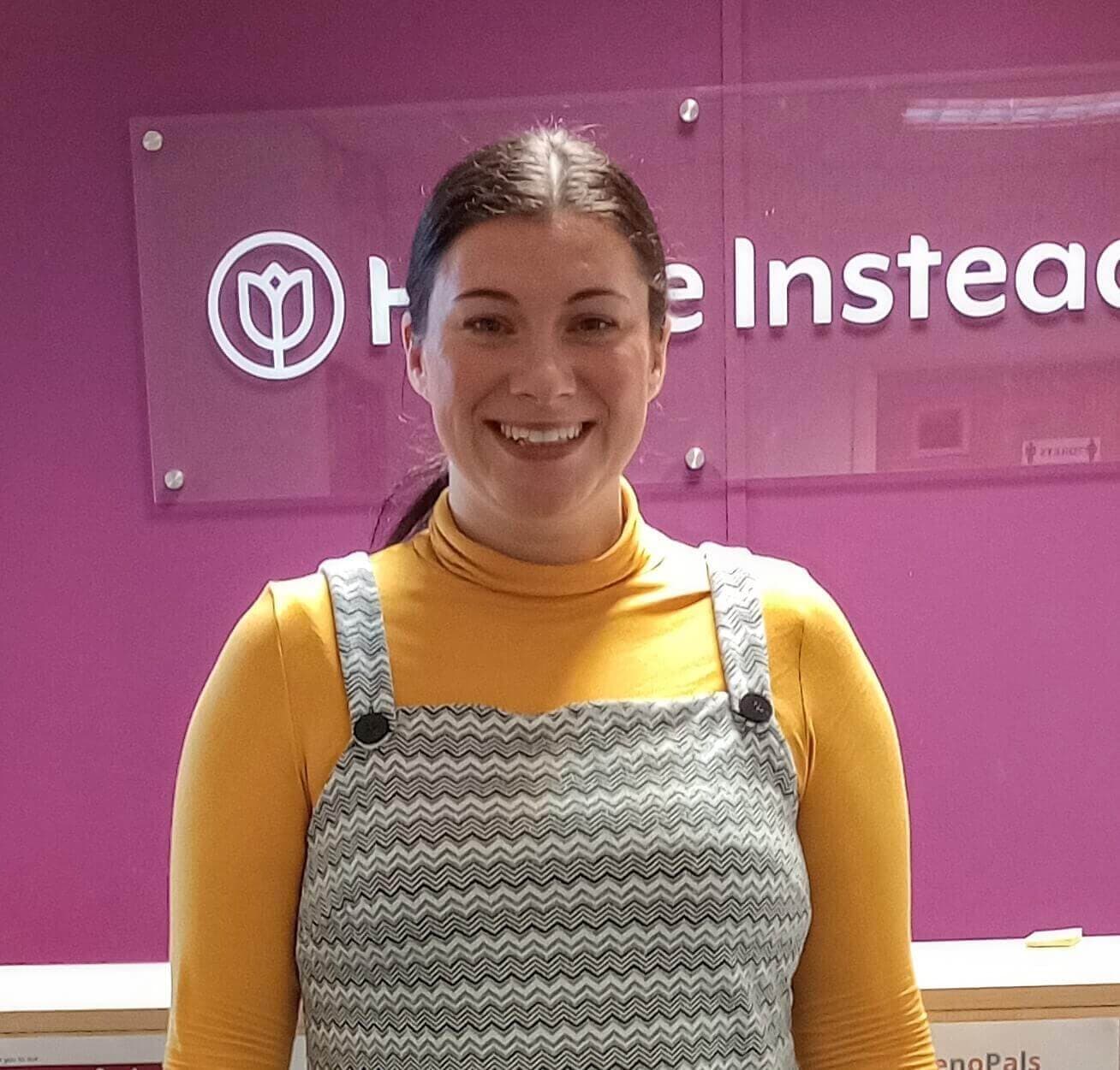 A woman in a yellow turtleneck and grey dress smiles in front of a purple wall with the "Home Instead" sign. - Home Instead