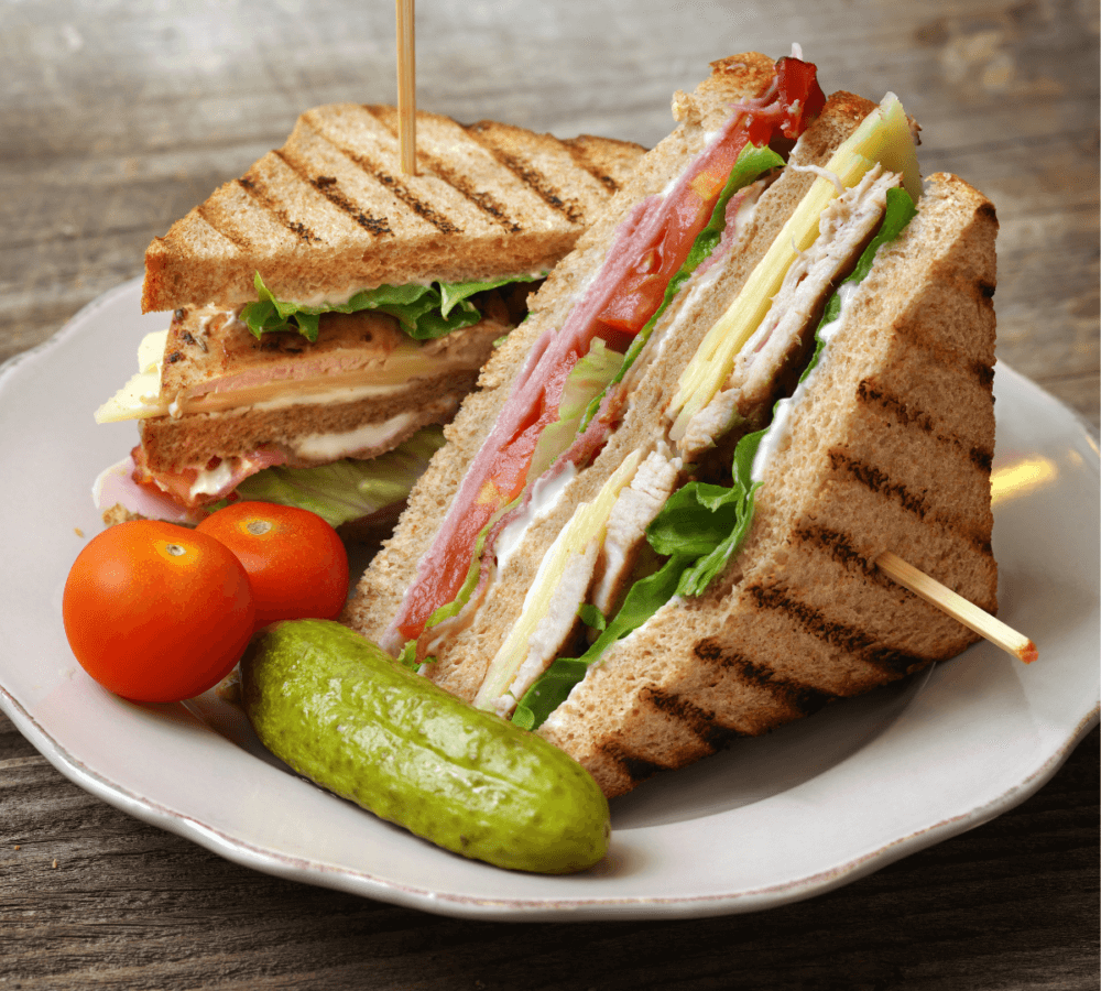 Club sandwich with layers of meats, cheese, lettuce, and tomato on a plate with cherry tomatoes and a pickle. - Home Instead