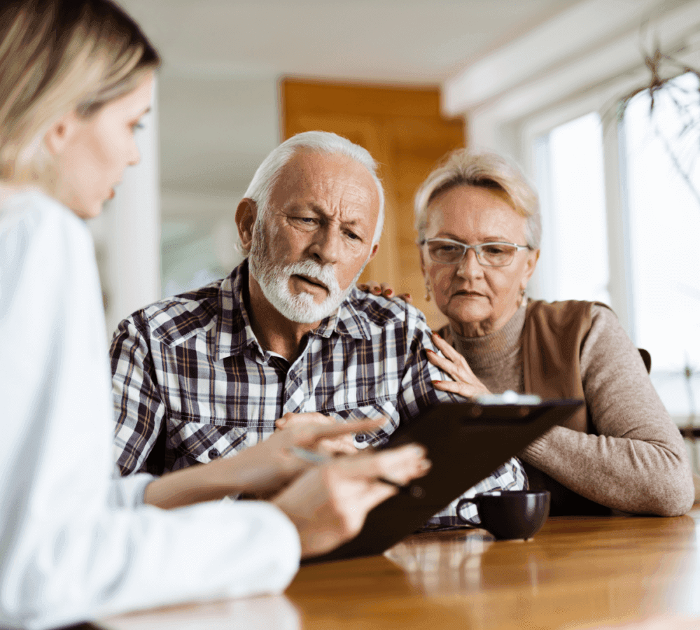 Elderly couple attentively listening to a young woman holding a clipboard, discussing paperwork at a table. - Home Instead