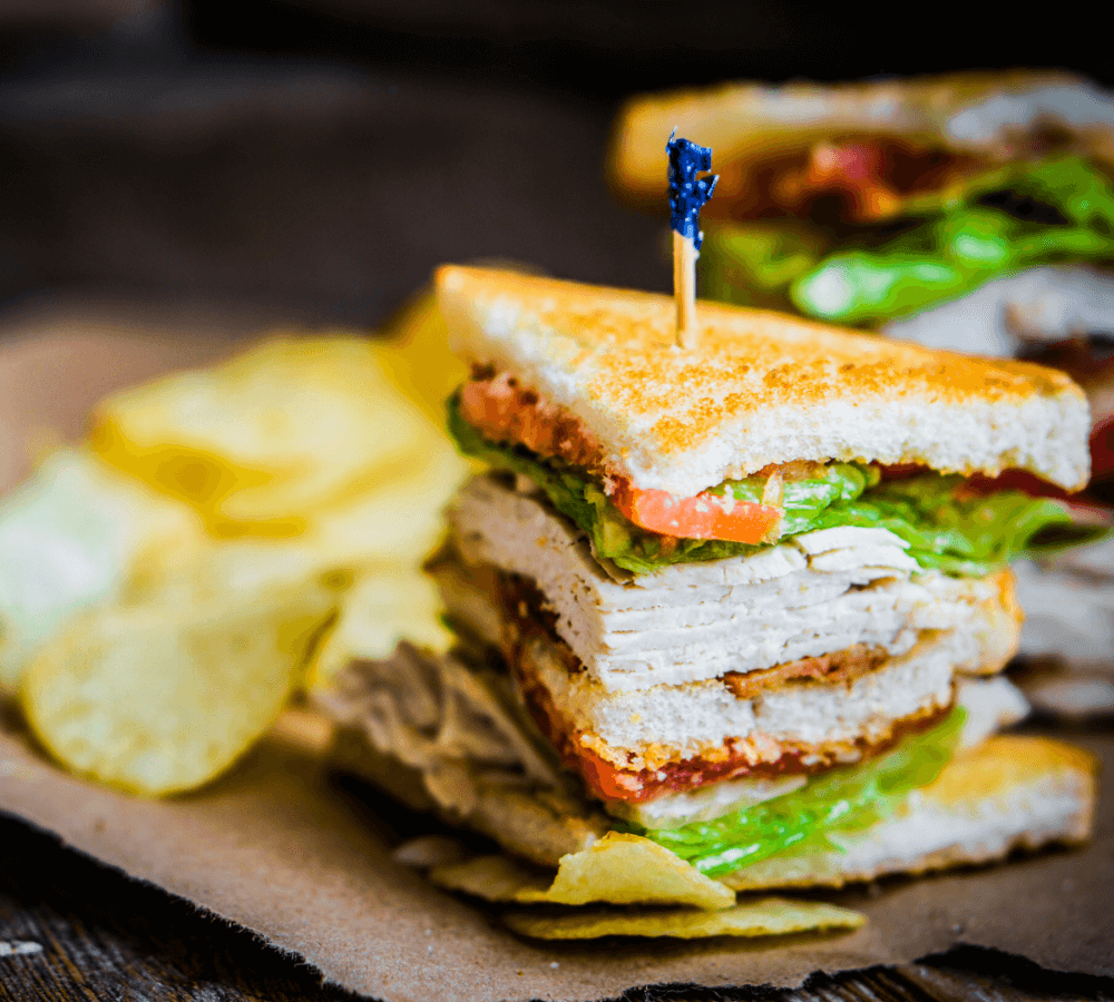 Close-up of a stacked turkey club sandwich with lettuce, tomato, and a toothpick, accompanied by potato chips. - Home Instead