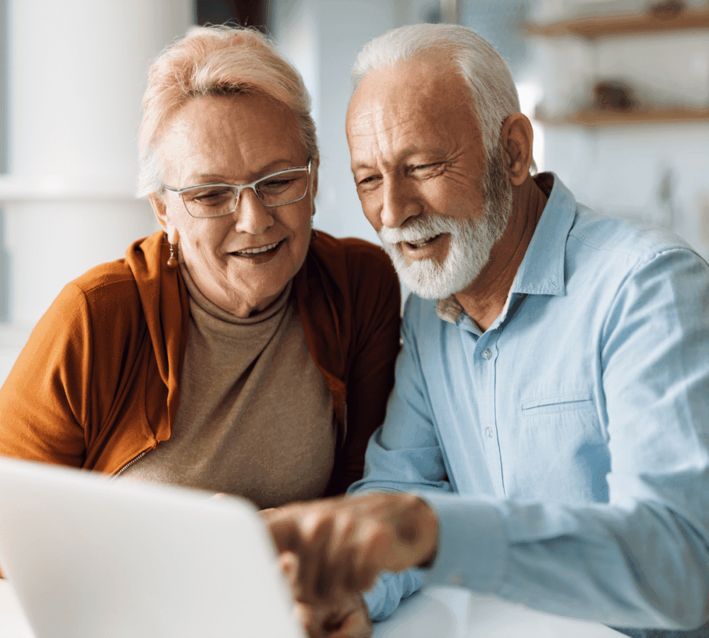 Two elderly people, smiling, look at a laptop screen together. The woman is wearing glasses. They seem engaged and happy. - Home Instead