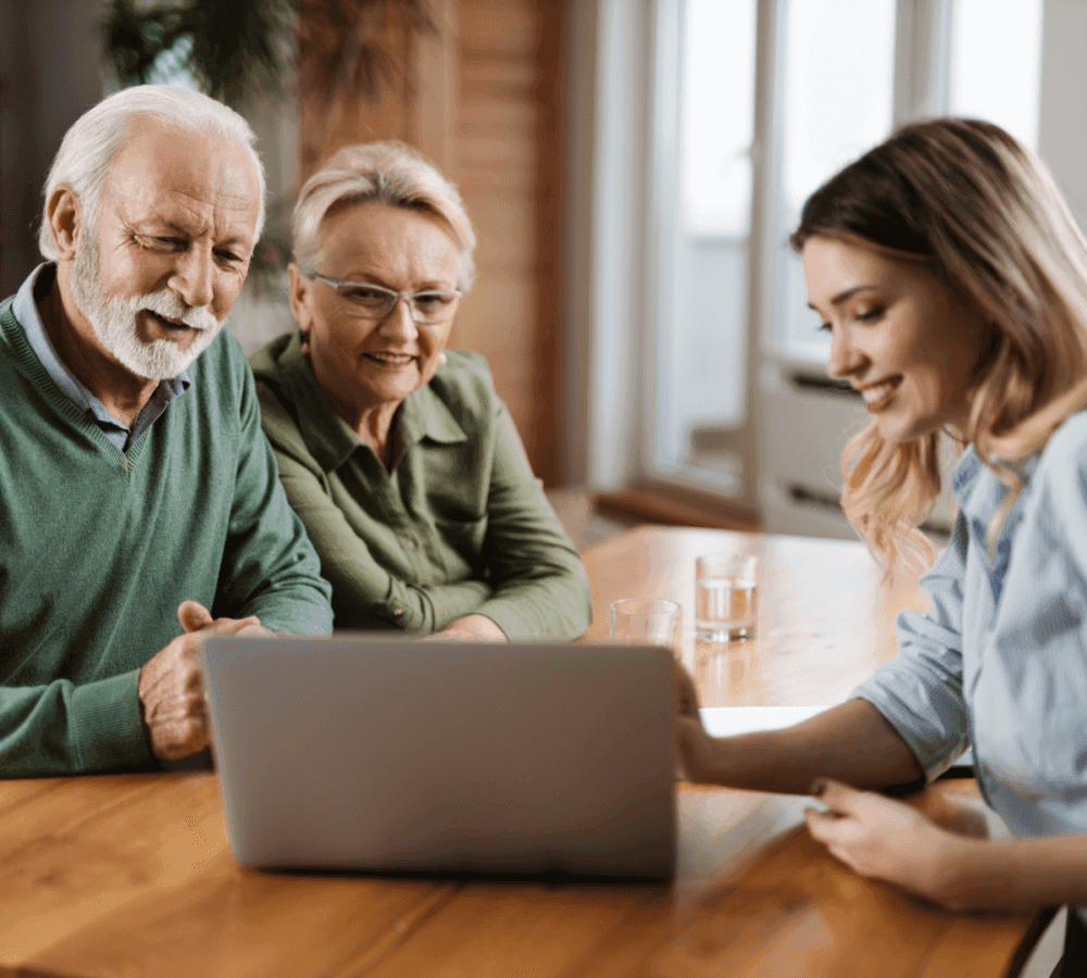 A senior couple smiles while a young woman shows them something on a laptop at a wooden table. - Home Instead