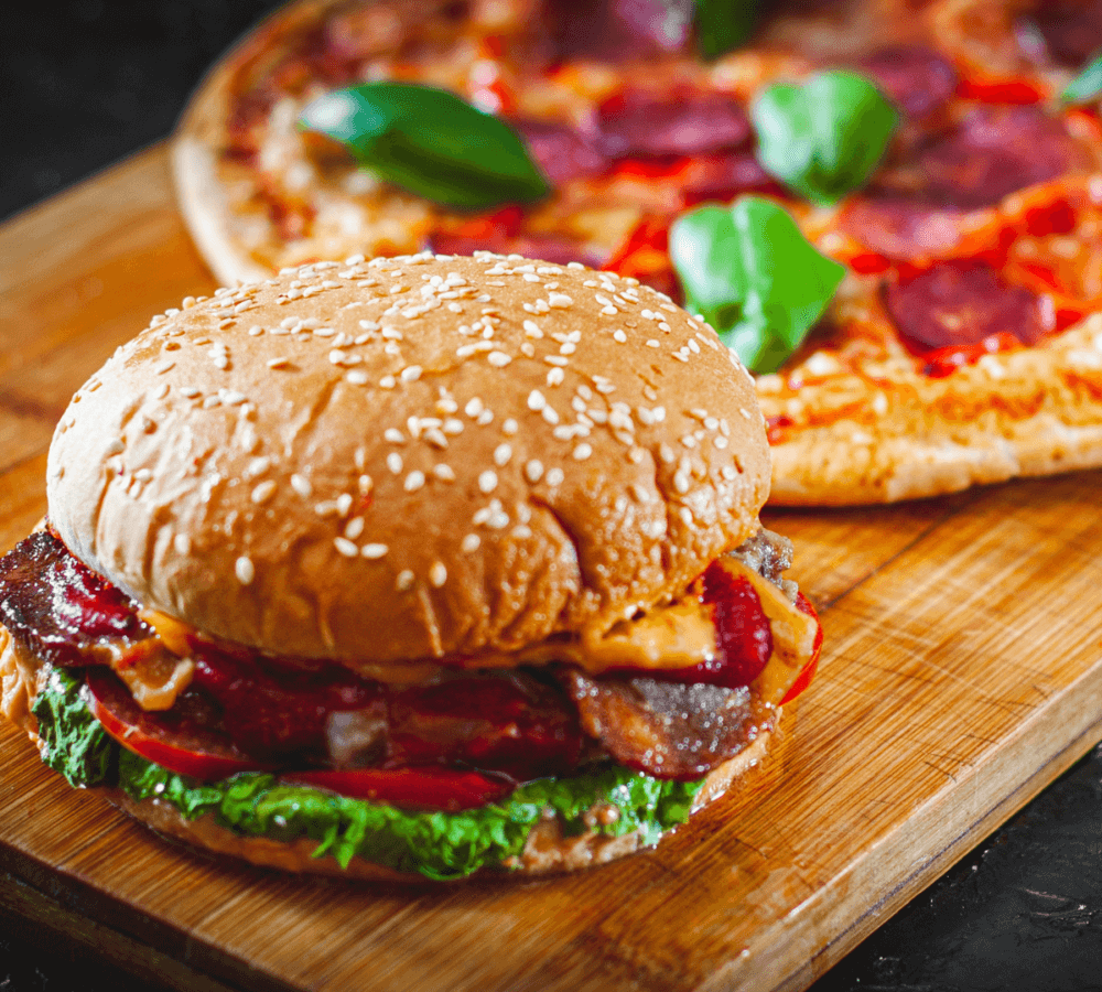 A sesame seed burger with lettuce and tomato on a wooden board, a pepperoni pizza in the background. - Home Instead