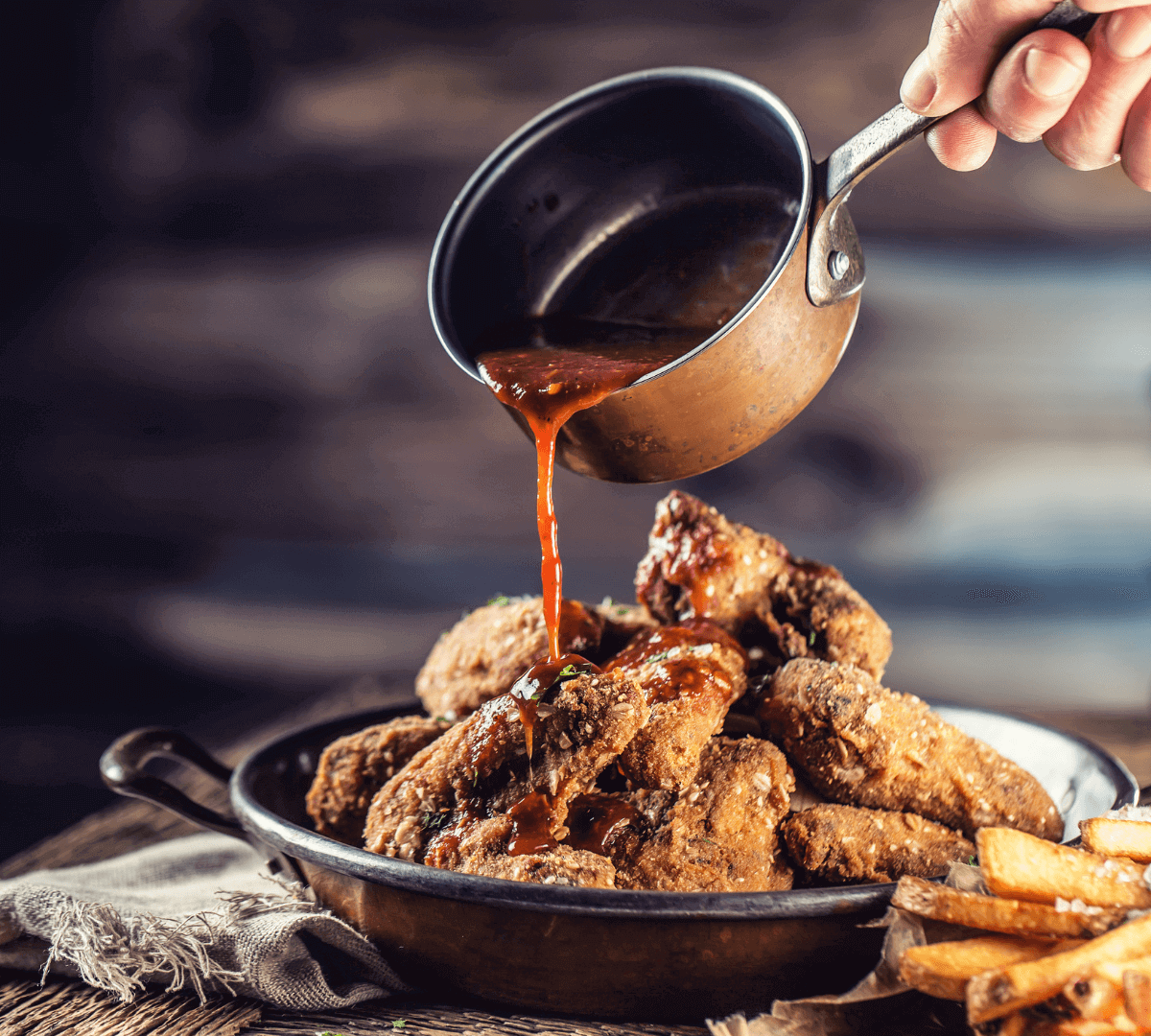 Hand pouring sauce over fried chicken wings in a rustic pan with a side of fries. - Home Instead