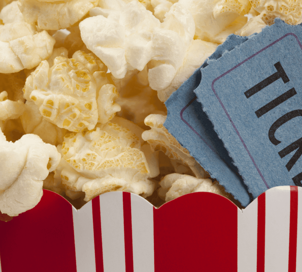 Close-up of popcorn in a red-striped container with two blue movie tickets tucked in. - Home Instead
