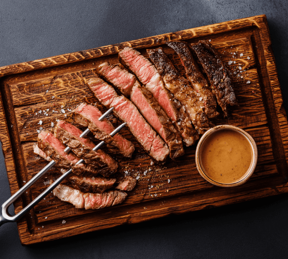 Sliced medium-rare steak on a wooden board with a side of sauce and a carving fork. - Home Instead