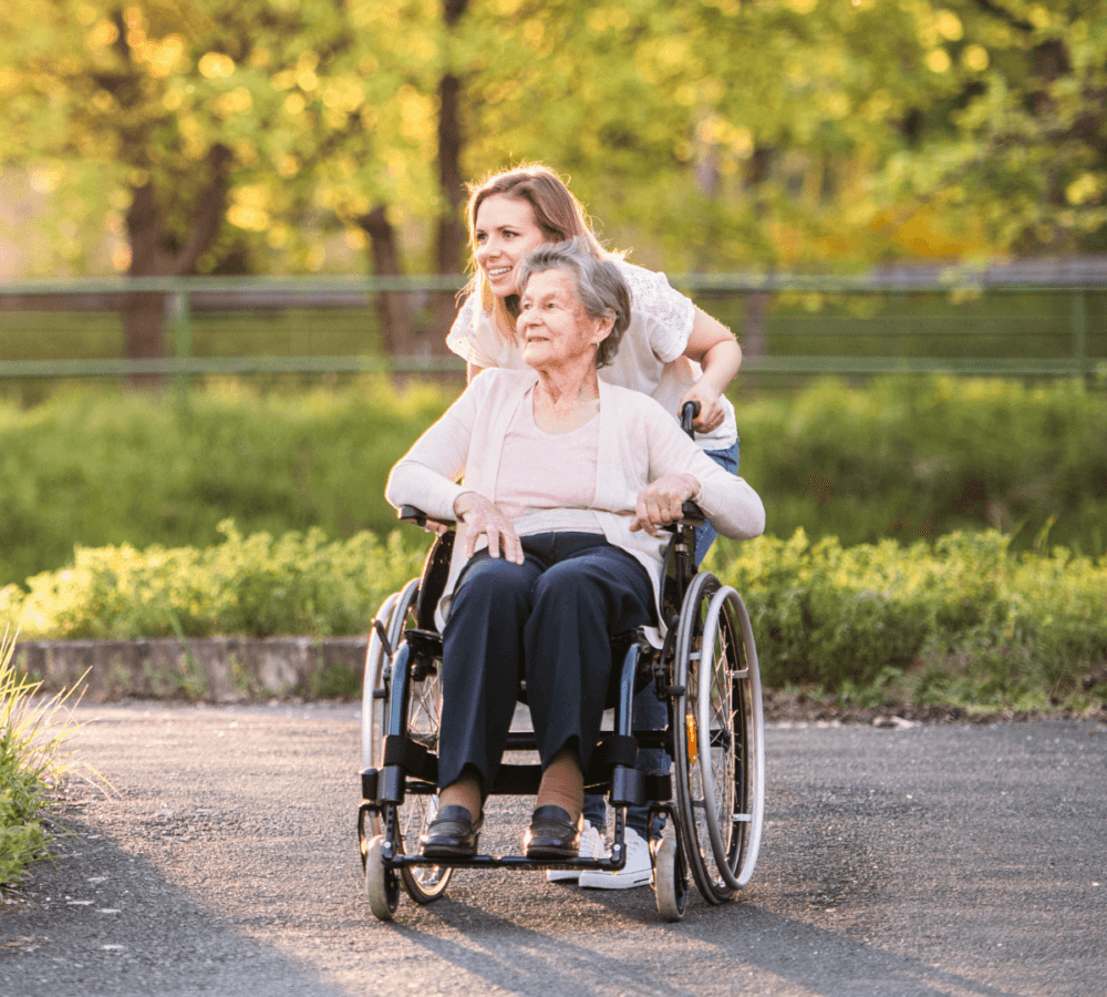 A woman pushes an elderly person in a wheelchair along a sunny path with green trees in the background. - Home Instead