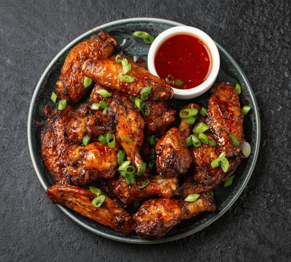 A plate of glazed chicken wings garnished with green onions, served with a side of red dipping sauce. - Home Instead