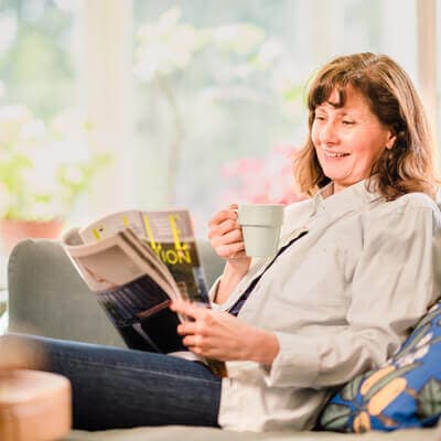 Woman sitting on a couch, smiling and drinking coffee while reading a magazine in a bright, cozy room. - Home Instead