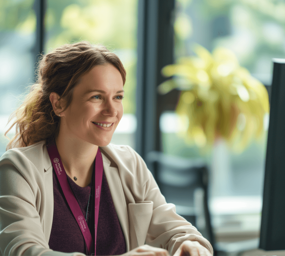 Smiling woman wearing a lanyard, sitting at a desk with a plant in the background. - Home Instead
