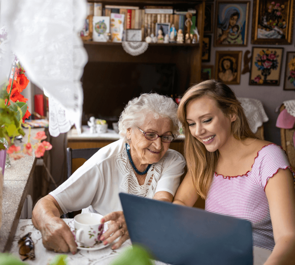 An elderly woman and a young woman are smiling and looking at a laptop screen together in a cozy, decorated room. - Home Instead