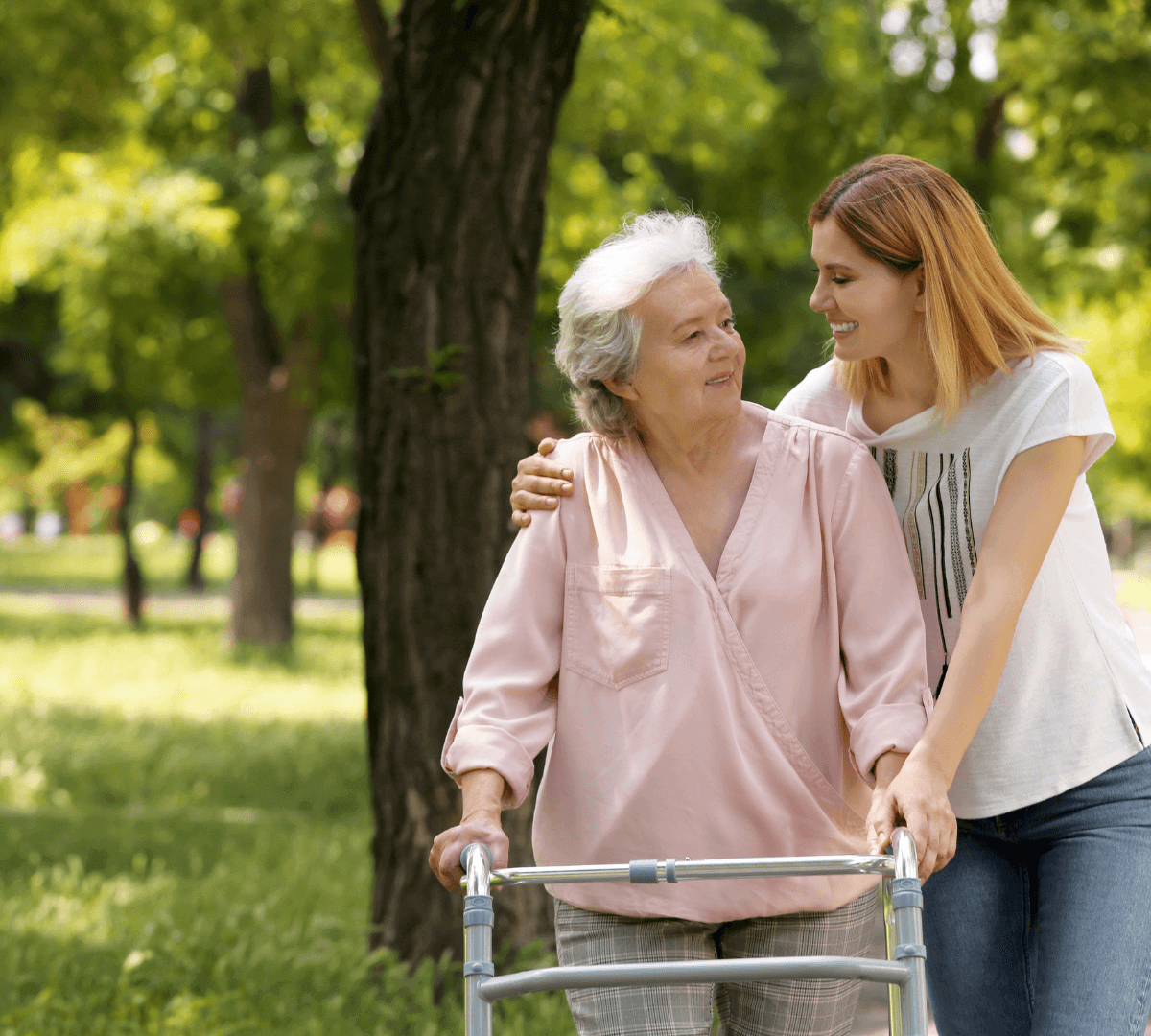 A young woman supports an elderly woman using a walker in a park, both smiling and sharing a moment. - Home Instead