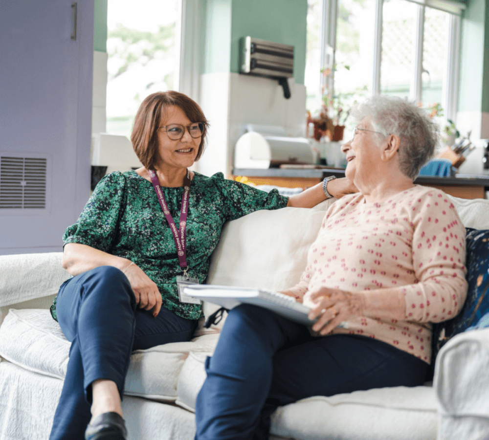 Two women smiling and talking while sitting on a sofa; one has a notepad on her lap. - Home Instead