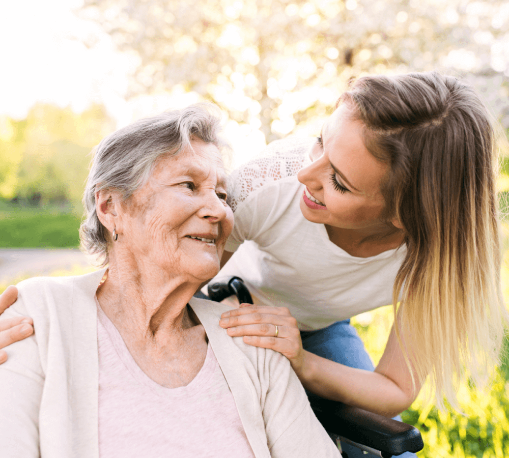 A young woman smiles at an elderly woman in a wheelchair outside in a sunny garden. - Home Instead