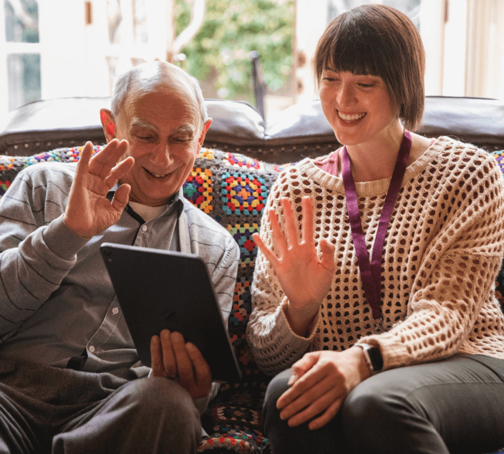 An elderly man and a young woman smiling and waving at a tablet while sitting on a colorful couch. - Home Instead
