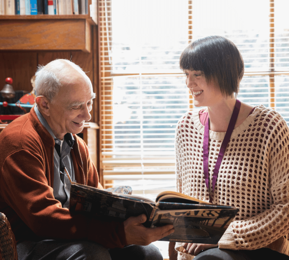 Elderly man and woman sit by a window, smiling and looking at a magazine together in a cozy room. - Home Instead