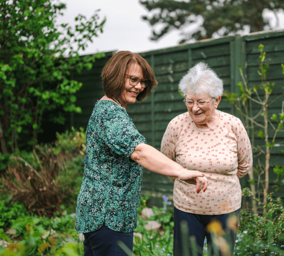 Two women in a garden, one pointing while the other smiles. Green fence and foliage in the background. - Home Instead