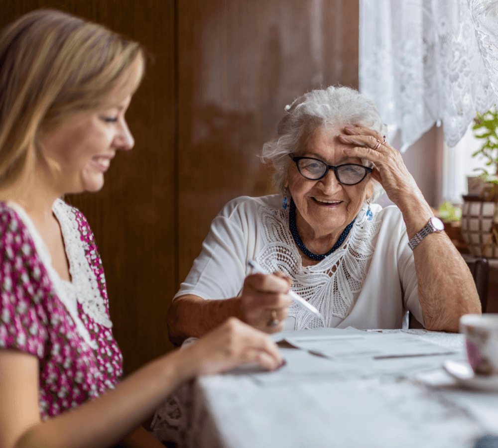 Elderly woman with glasses smiling and holding a pen, sitting next to a younger woman at a table. - Home Instead