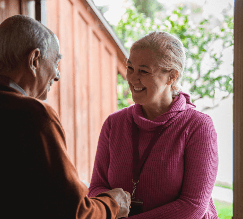 Two elderly people smiling and shaking hands in a doorway, with one person wearing a pink sweater and a lanyard. - Home Instead