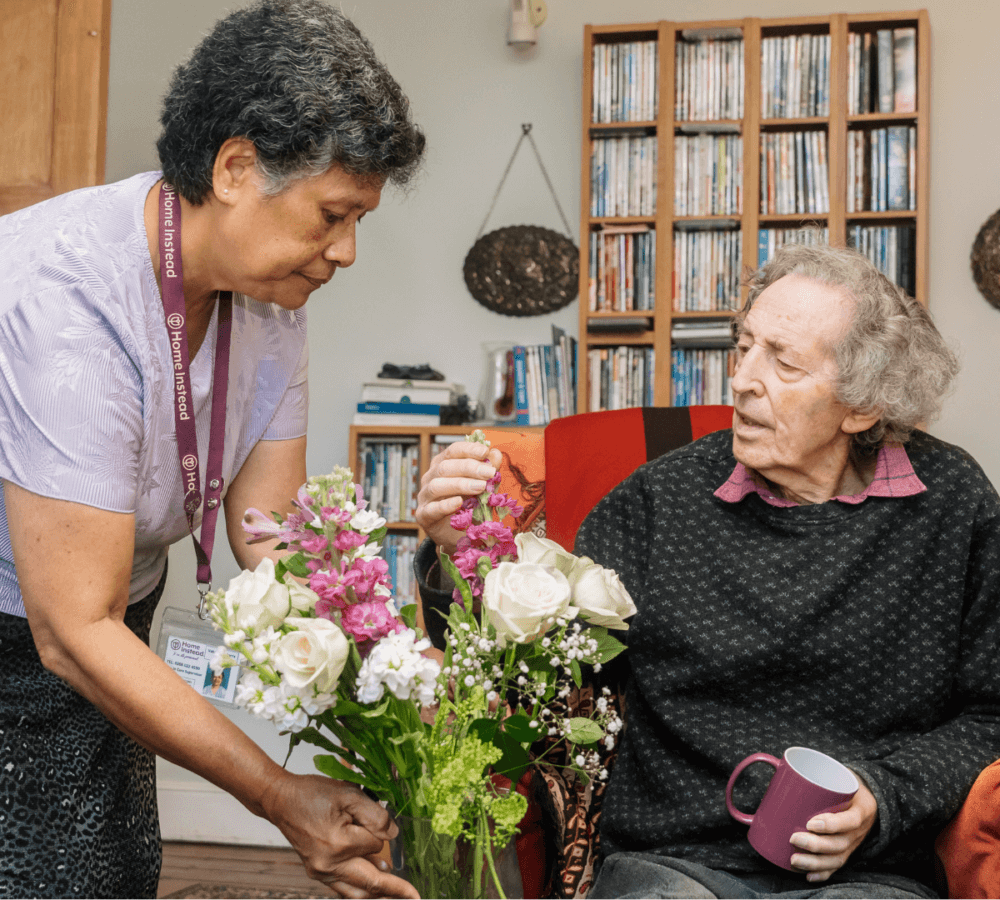 A caregiver helps an elderly person arrange a bouquet of flowers while sitting in a cozy room with bookshelves. - Home Instead