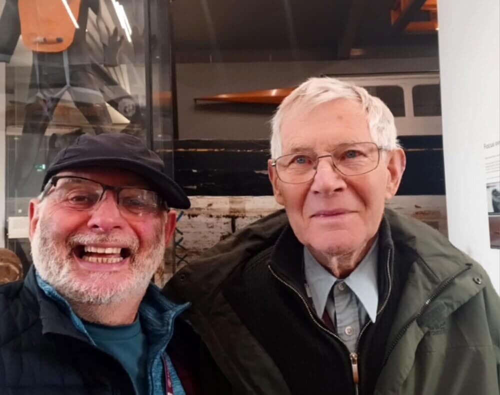 Two elderly men smiling, one wearing a cap and glasses, the other with white hair and glasses, indoors. - Home Instead
