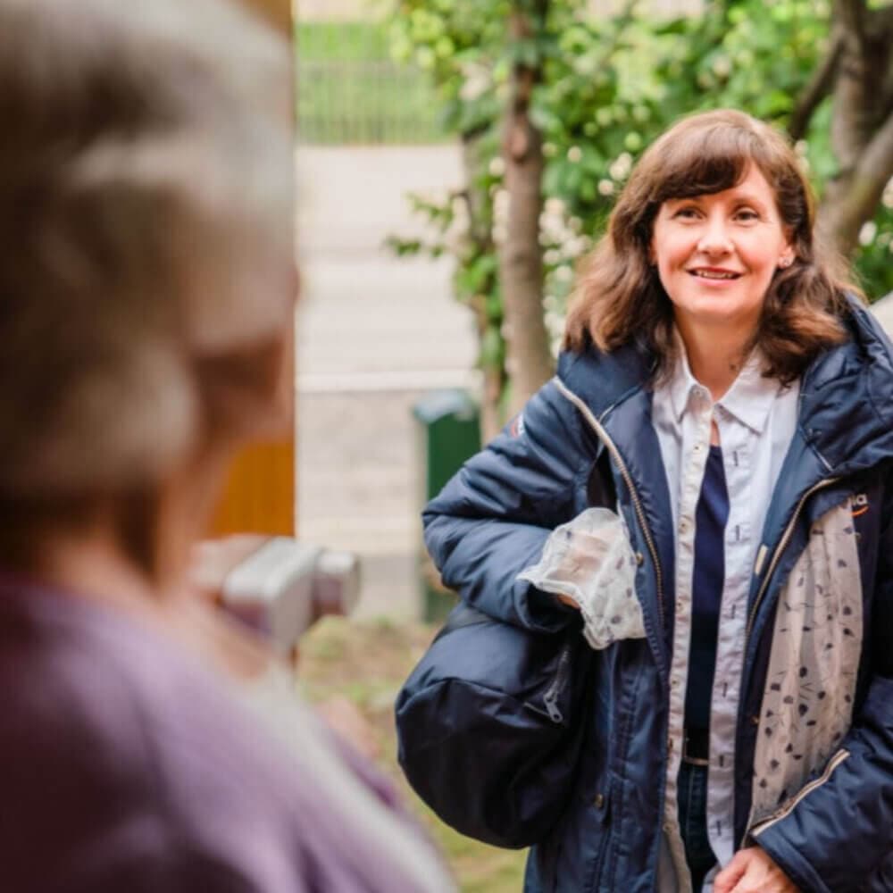 A woman holding a bag smiles and greets an older person at the door on a sunny day. - Home Instead