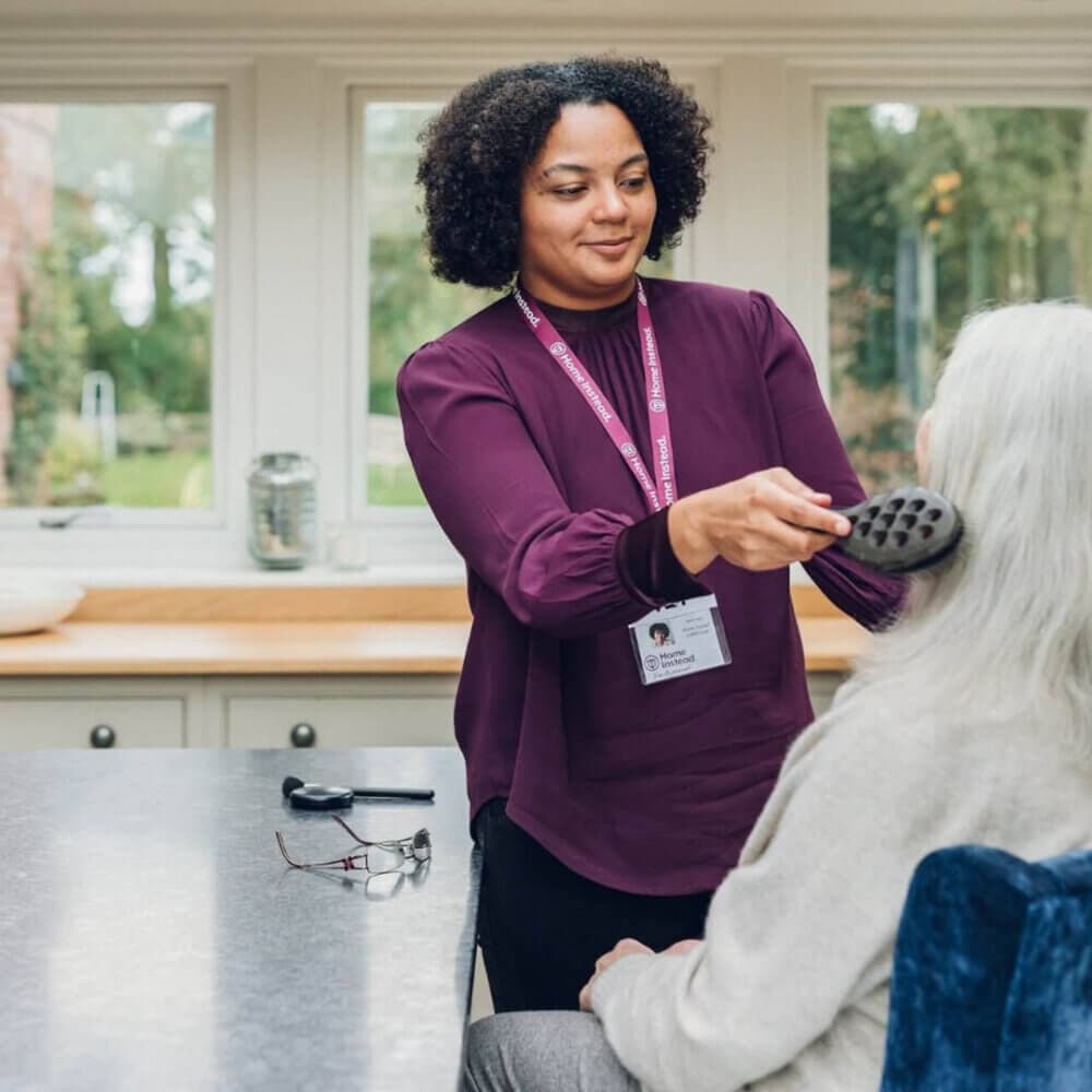 A caregiver with a lanyard brushes an elderly person's hair in a bright room with large windows. - Home Instead