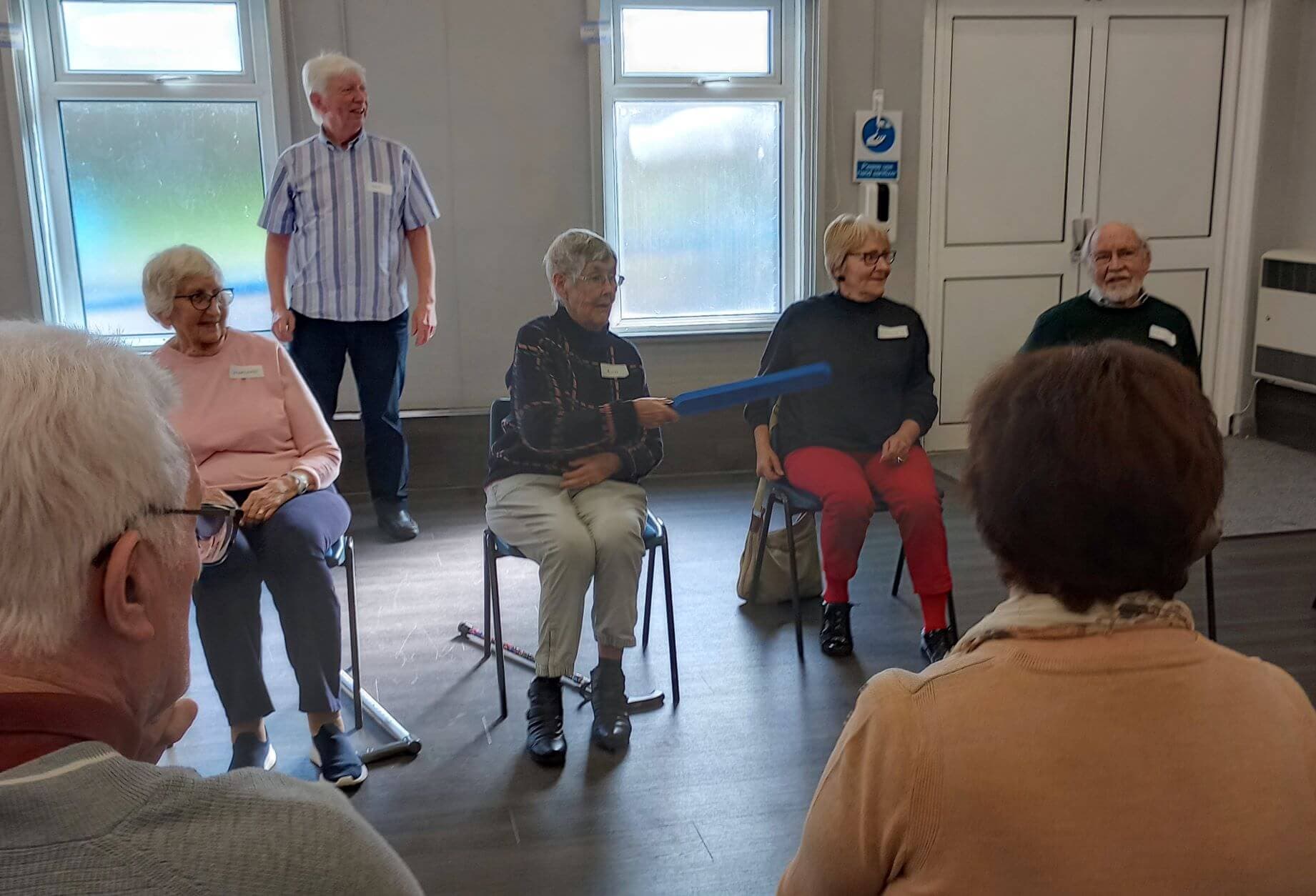 A group of elderly people sit in a circle participating in an indoor activity. One holds a blue foam baton. - Home Instead